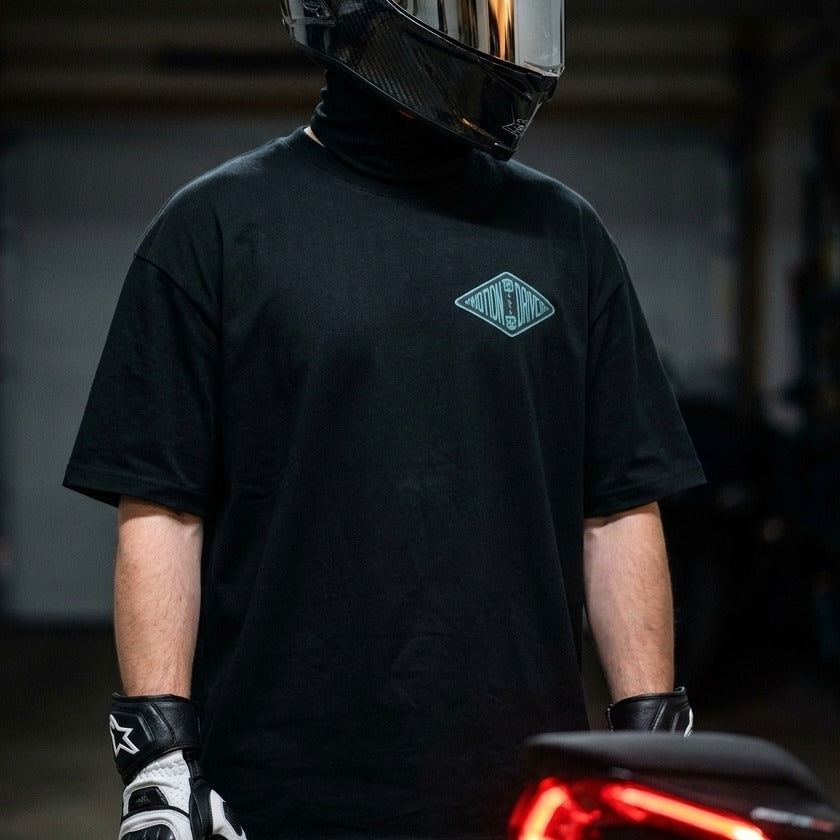 A person wearing a black short-sleeve t-shirt with a small diamond-shaped teal graphic on the left chest, paired with a black neck gaiter, black and white gloves, and a carbon-fiber style full-face helmet in a dark indoor setting.