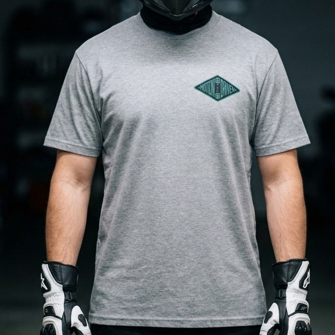 A person wearing a heather gray short-sleeve t-shirt with a small green diamond-shaped chest graphic that reads 'THE WAY'. The person is also wearing black and white motorcycle gloves and a black face mask, set against a dark, blurred background.