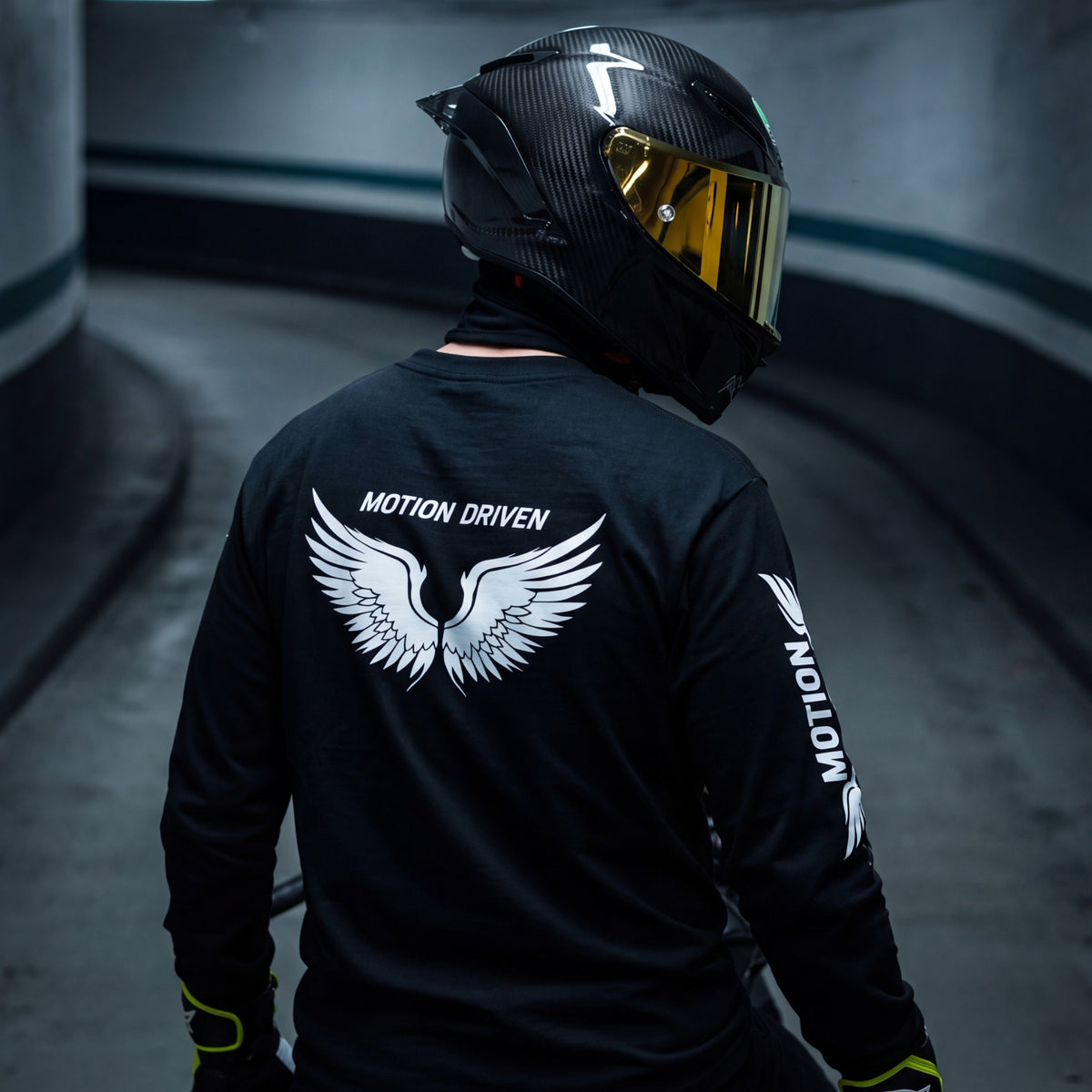 A person wearing a black long-sleeve racing shirt and a black carbon-fiber motorcycle helmet with a gold visor, photographed from behind in a curved tunnel. The shirt features a large white wing graphic and the text 'MOTION DRIVEN' on the back, with 'MOTION' printed down the sleeve.