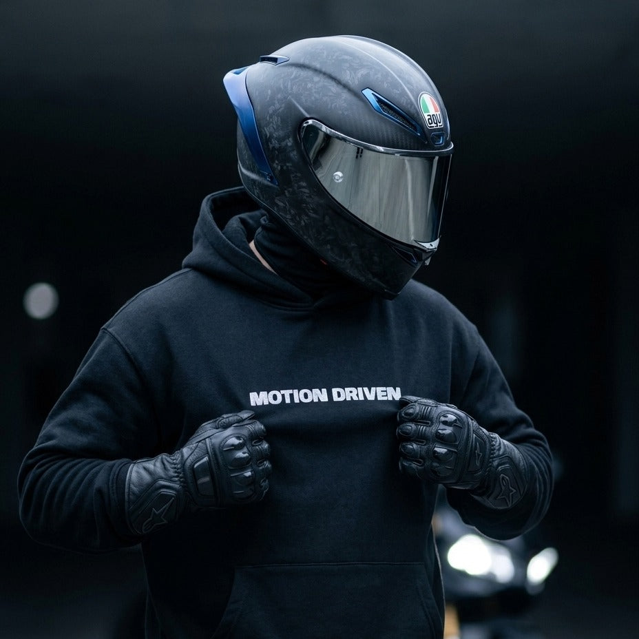 A person wearing a black full-face motorcycle helmet with a clear visor, black gloves, and a black hoodie with the text 'MOTION DRIVEN' across the chest, posed against a dark blurred background.