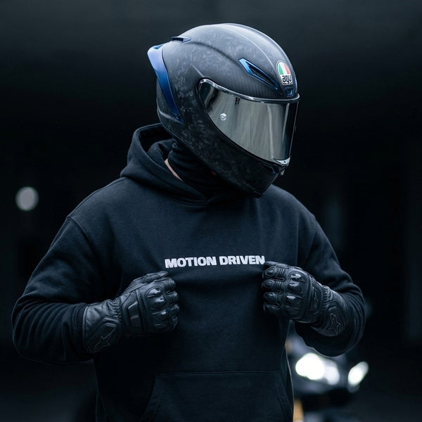 A person wearing a black full-face motorcycle helmet with a clear visor, black gloves, and a black hoodie with the text 'MOTION DRIVEN' across the chest, posed against a dark blurred background.