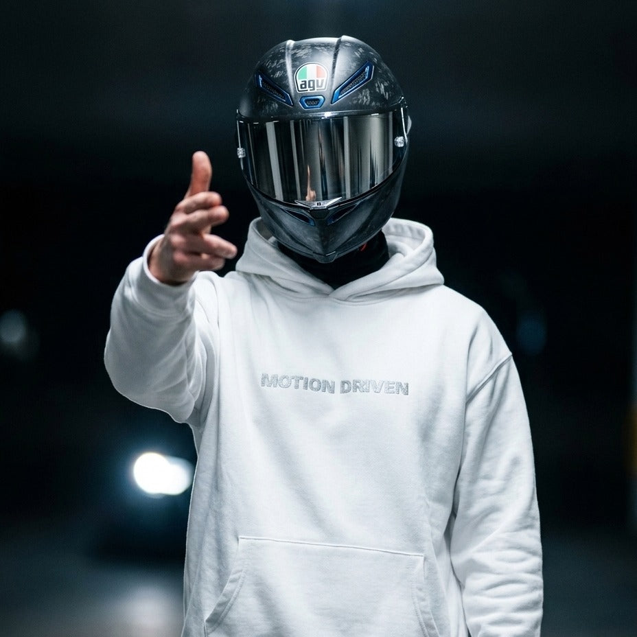 A person wearing a black AGV full-face motorcycle helmet and a white hoodie with 'MOTION DRIVEN' printed on the chest, pointing forward against a dark blurred background.