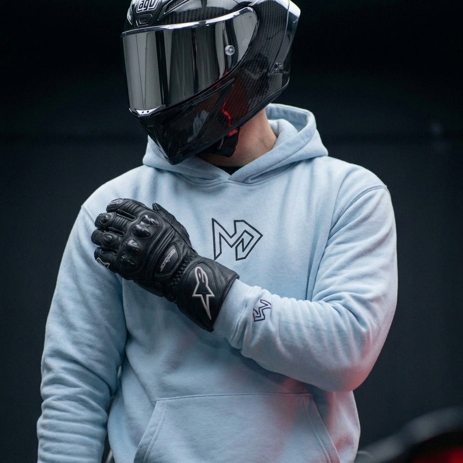 A person wearing a light blue hoodie with a black graphic 'M' on the chest, black motorcycle gloves, and a black full-face helmet, posed against a dark background.