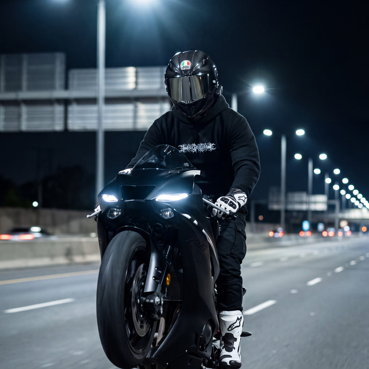 A person wearing a black full-face helmet, black hoodie, black pants, and white-and-black gloves rides a black sport motorcycle on a highway at night under streetlights.