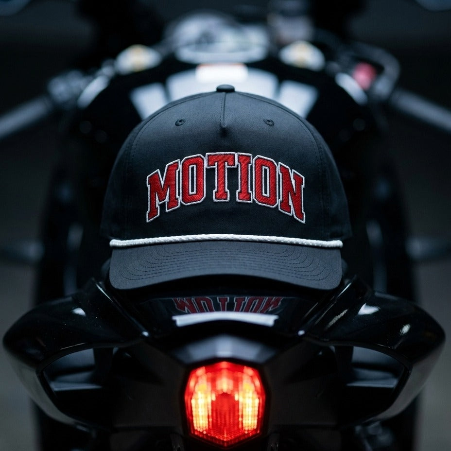 A black baseball cap with 'MOTION' embroidered in red arched letters and a white rope detail on the brim, resting on the back of a black motorcycle.