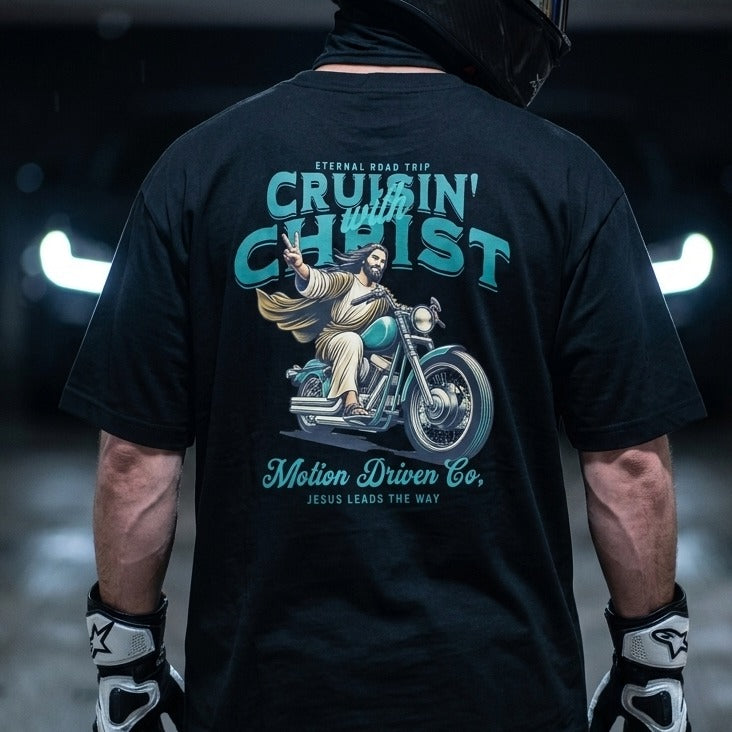A person shown from the back wearing a black graphic T-shirt. The shirt features a large teal and beige print of Jesus riding a motorcycle with the text 'CRUISE WITH CHRIST' and 'Motion Driven Co.