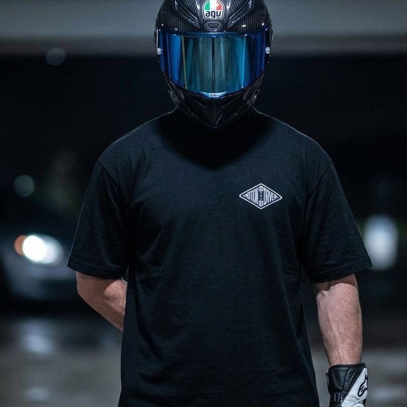 A person wearing a black full-face motorcycle helmet with a reflective visor and a black short-sleeve T-shirt with a small white chest logo, standing in a dark outdoor setting.