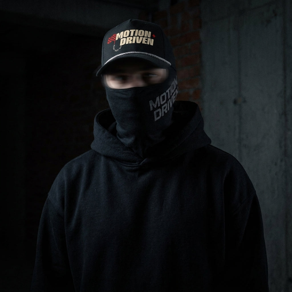 A person wearing a black hoodie and a black face covering with 'MOTION DRIVEN' printed on it, also wearing a black cap with the same logo, set against a dark concrete background.