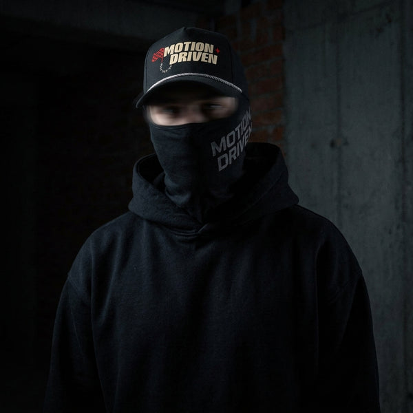 A person wearing a black hoodie and a black face covering with 'MOTION DRIVEN' printed on it, also wearing a black cap with the same logo, set against a dark concrete background.