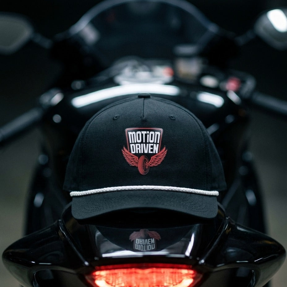 A black baseball cap with a white rope detail and a red embroidered winged logo patch reading 'MOTION DRIVEN' sits on the back of a black motorcycle in a dark setting.