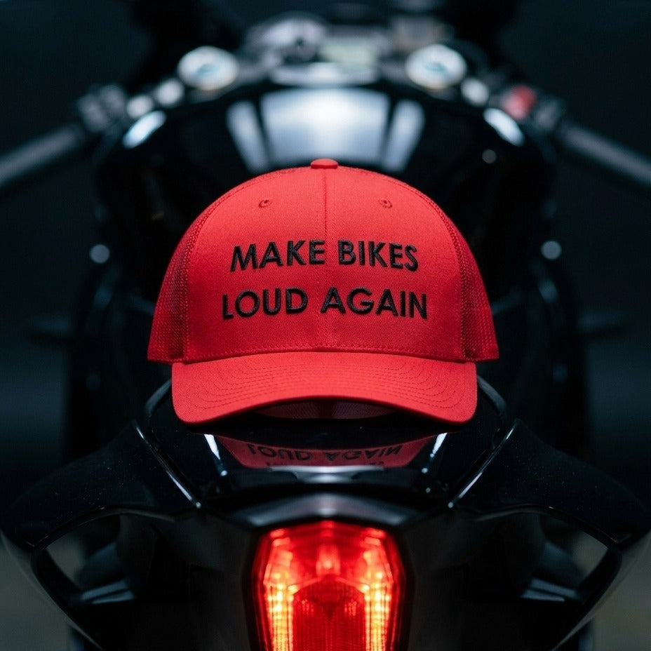 A red baseball cap with black embroidered text reading 'MAKE BIKES LOUD AGAIN' sits on the back of a black motorcycle, with the motorcycle's taillight glowing in the background.
