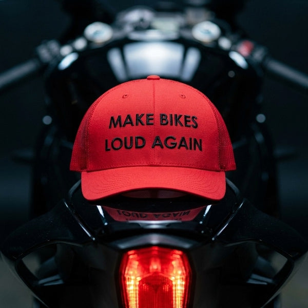 A red baseball cap with black embroidered text reading 'MAKE BIKES LOUD AGAIN' sits on the back of a black motorcycle, with the motorcycle's taillight glowing in the background.