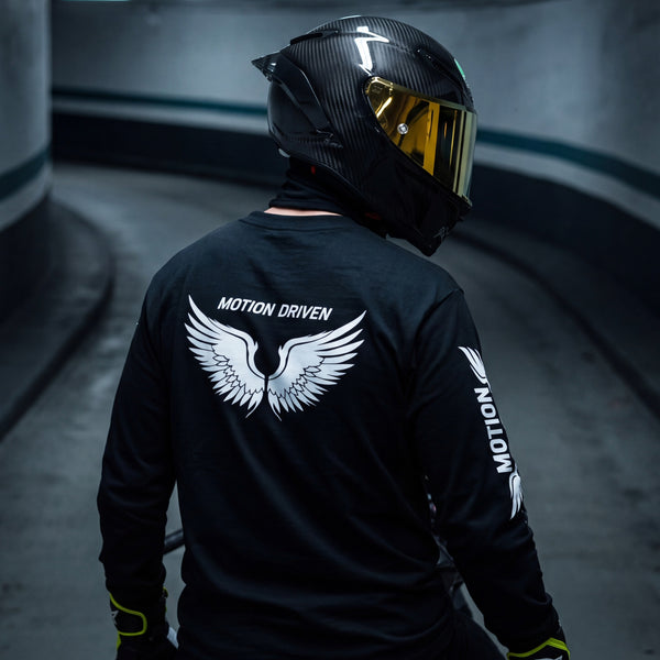 A person wearing a black long-sleeve racing shirt and a black carbon-fiber motorcycle helmet with a gold visor, photographed from behind in a curved tunnel. The shirt features a large white wing graphic and the text 'MOTION DRIVEN' on the back, with 'MOTION' printed down the sleeve.