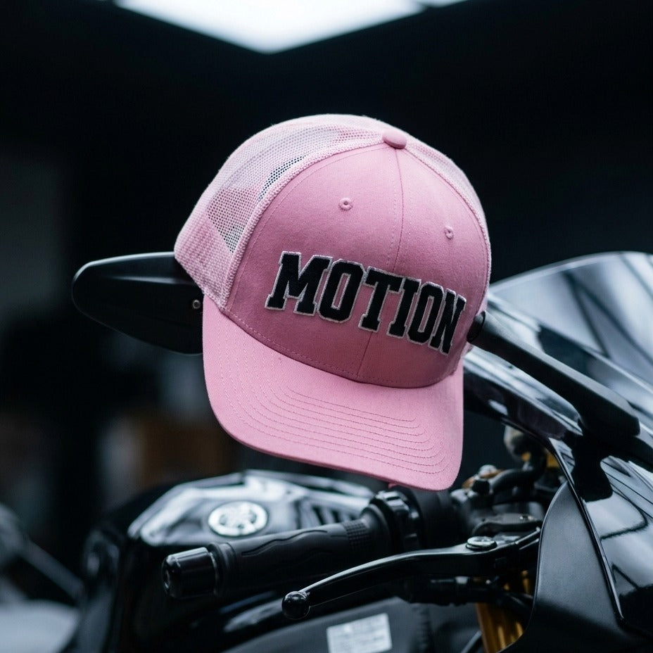 A pink trucker-style cap with black 'MOTION' lettering on the front, resting on the handlebars of a black motorcycle against a dark blurred background.