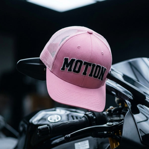 A pink trucker-style cap with black 'MOTION' lettering on the front, resting on the handlebars of a black motorcycle against a dark blurred background.