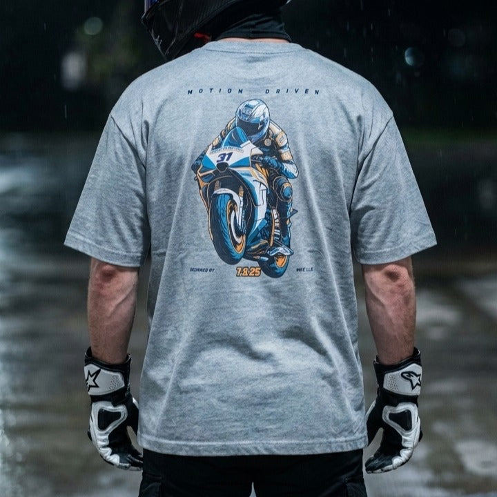 A person shown from behind wearing a light gray t-shirt with a large blue and white motorcycle graphic on the back, along with black and white racing gloves, set against a dark, rainy outdoor background.