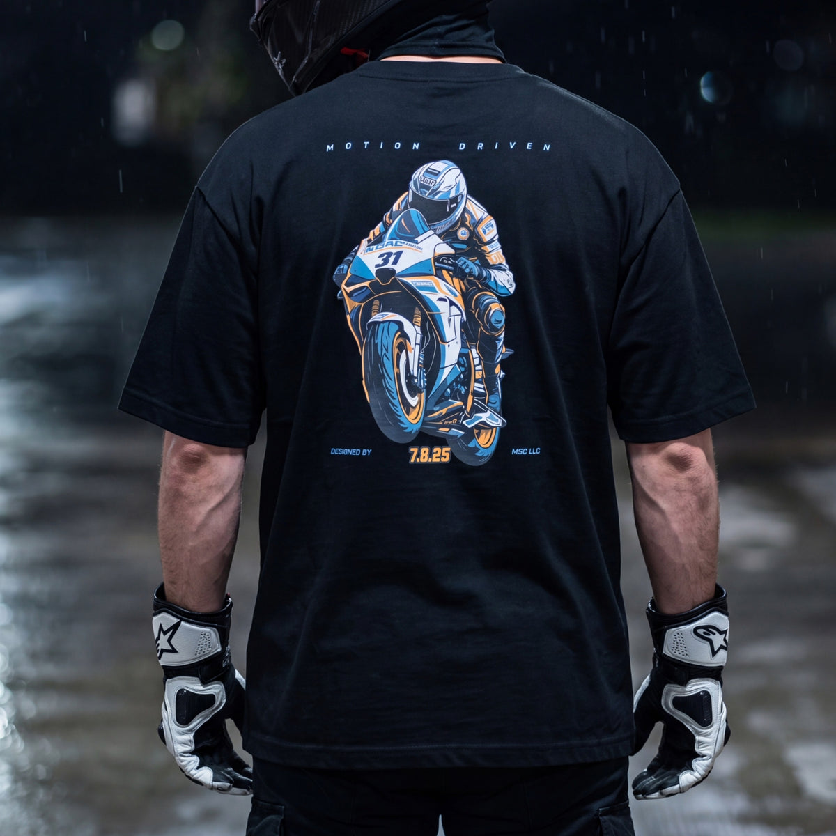 A person shown from behind wearing a black graphic T-shirt with a large motorcycle racer print on the back, black gloves, and a dark hood. The background is a blurred rainy street scene.