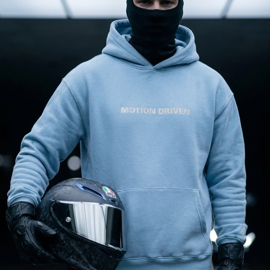 A person wearing a light blue hoodie with 'MOTION DRIVEN' printed on the chest, black gloves, and a black face covering, holding a black motorcycle helmet with a reflective visor.