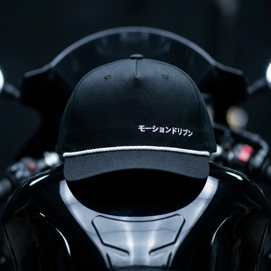 A black baseball cap with white embroidered Japanese text and a white rope detail on the brim, displayed on a glossy black motorcycle helmet against a dark blurred background.