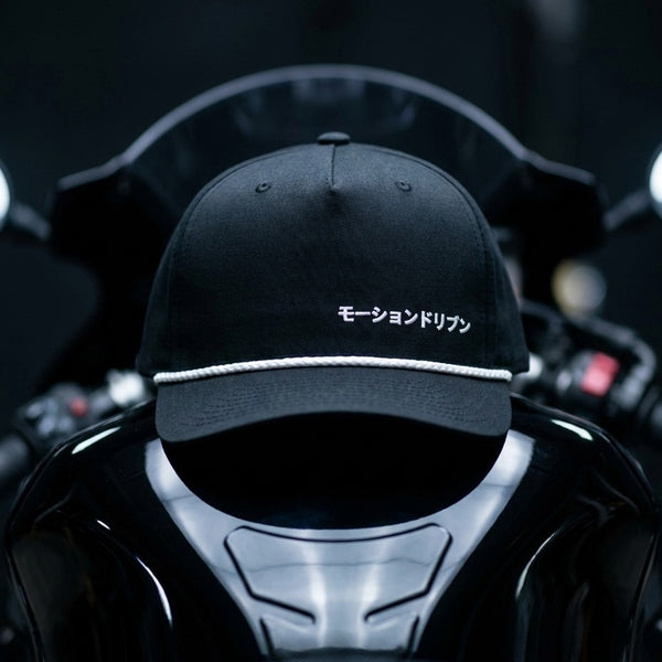A black baseball cap with white embroidered Japanese text and a white rope detail on the brim, displayed on a glossy black motorcycle helmet against a dark blurred background.