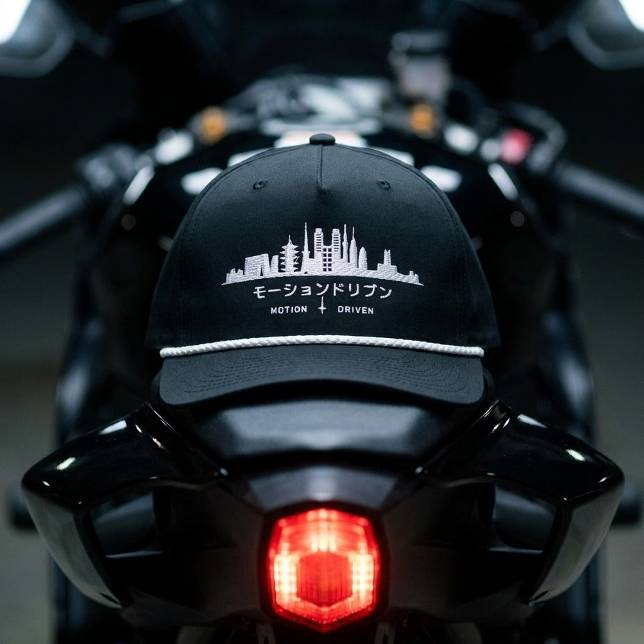 A black baseball cap with a white embroidered motorcycle cityscape and the text 'モーションドリブン MOTION + DRIVEN' resting on the back of a black motorcycle.