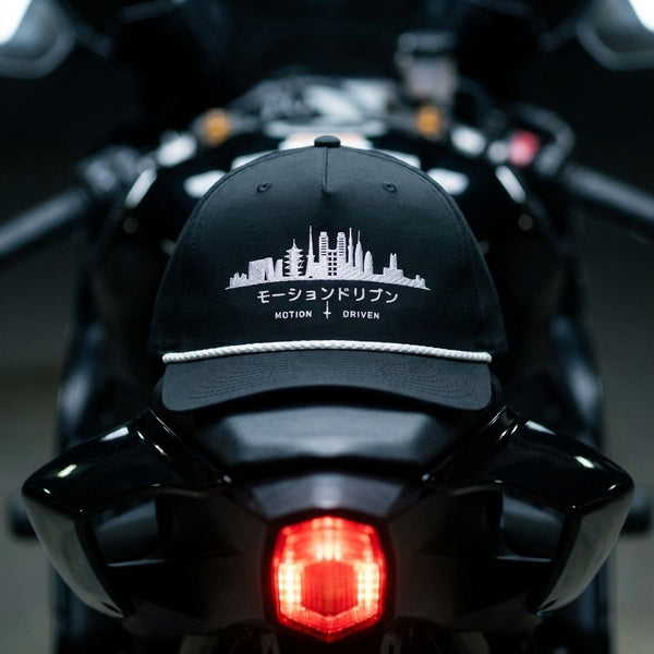 A black baseball cap with a white embroidered motorcycle cityscape and the text 'モーションドリブン MOTION + DRIVEN' resting on the back of a black motorcycle.
