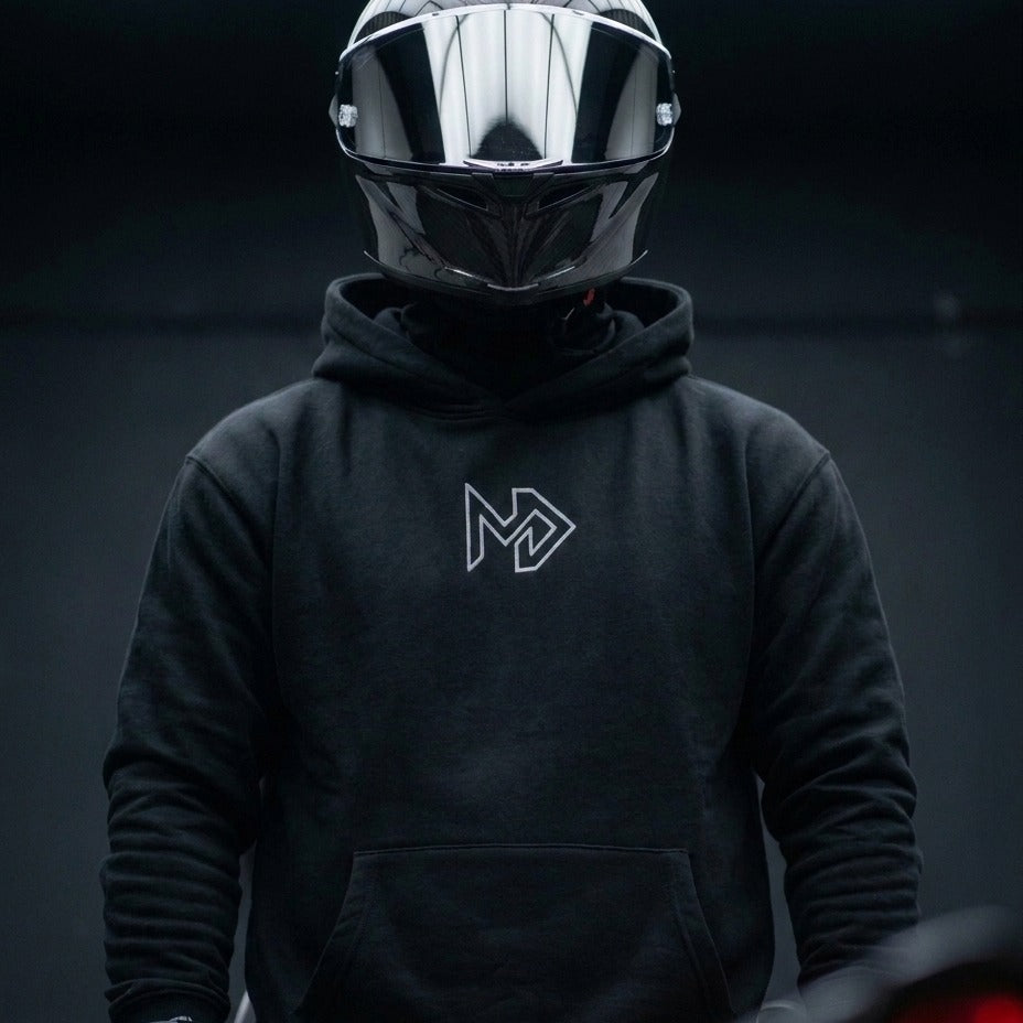 A person wearing a glossy black full-face motorcycle helmet and a black pullover hoodie with a white stylized 'MD' logo on the chest, set against a dark, moody background.