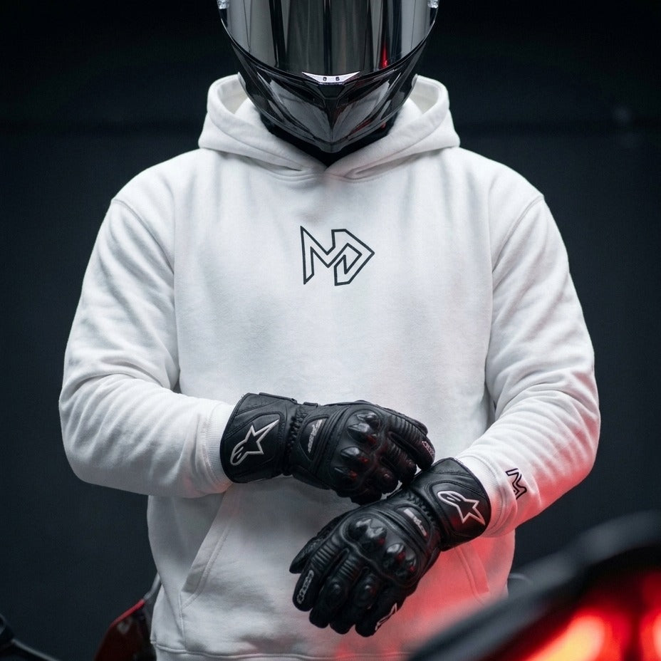 A person wearing a white pullover hoodie with a black geometric logo on the chest, black motorcycle gloves, and a black full-face helmet, standing against a dark background.
