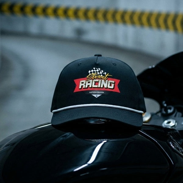 A black baseball cap with a 'Street Racing' graphic on the front, resting on the glossy tank of a black motorcycle in a blurred outdoor setting.