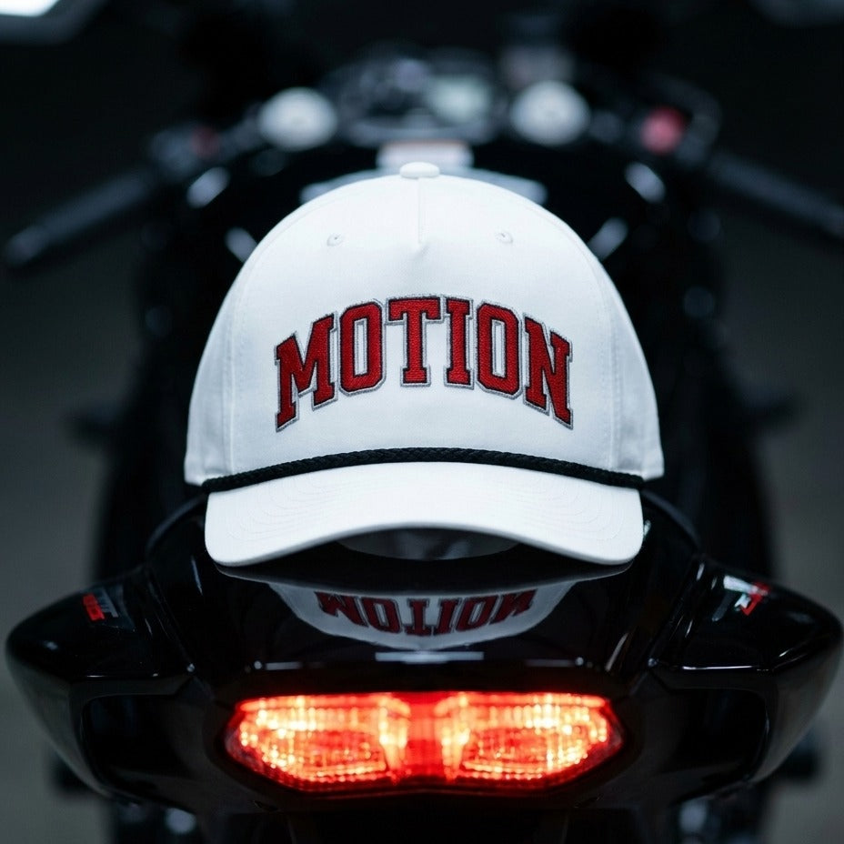 A white baseball cap with 'MOTION' embroidered in red across the front, resting on the glossy black rear of a motorcycle with its red taillight illuminated.