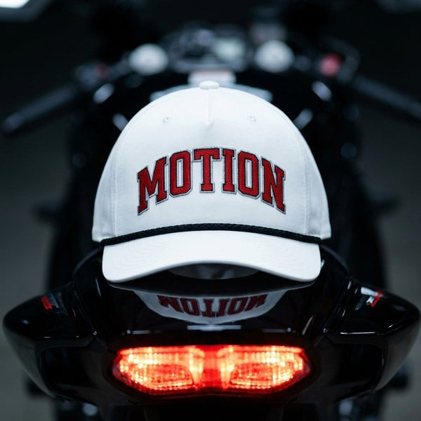 A white baseball cap with 'MOTION' embroidered in red across the front, resting on the glossy black rear of a motorcycle with its red taillight illuminated.